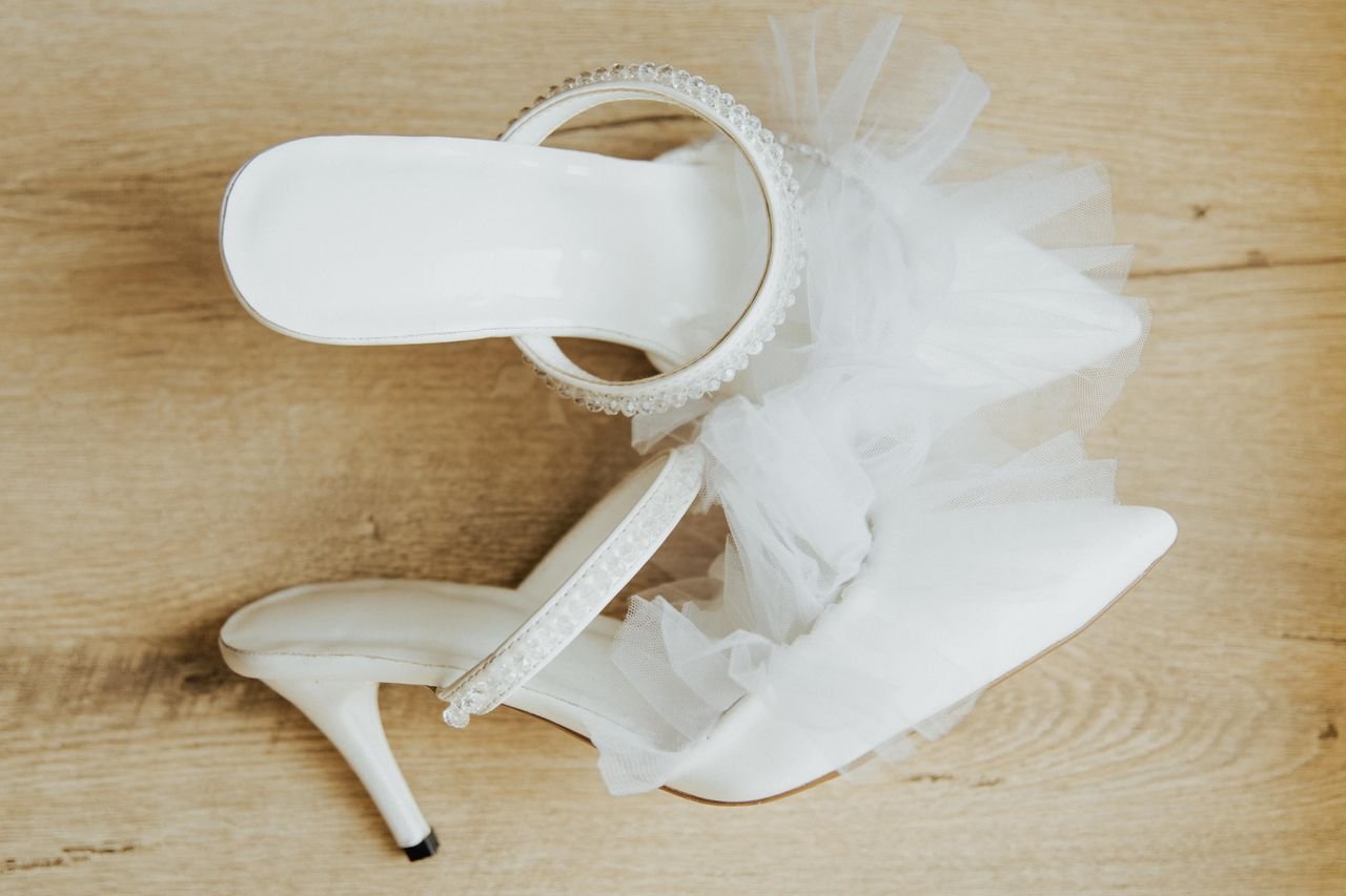About A stylish pair of white bridal high heel shoes with tulle detail, elegantly posed on a wooden floor.